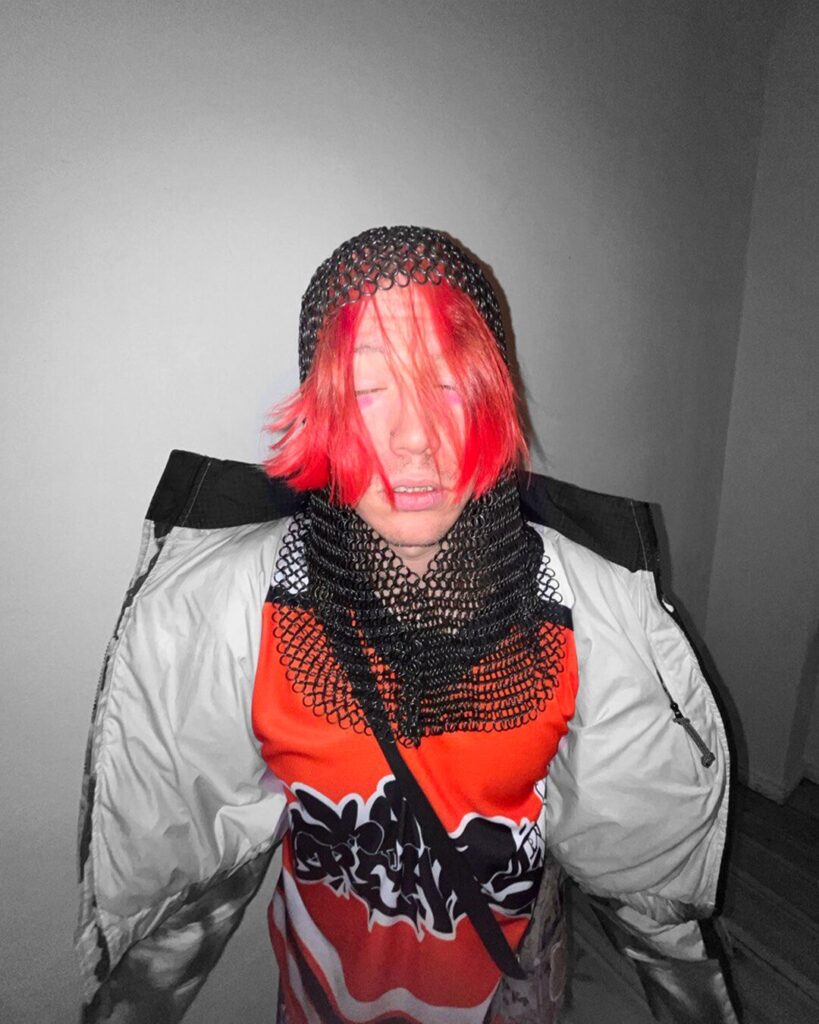 Person mit roten Haaren trägt ein schwarzes Kettenhaube über einem roten Shirt mit schwarzem Muster und eine offene helle Jacke