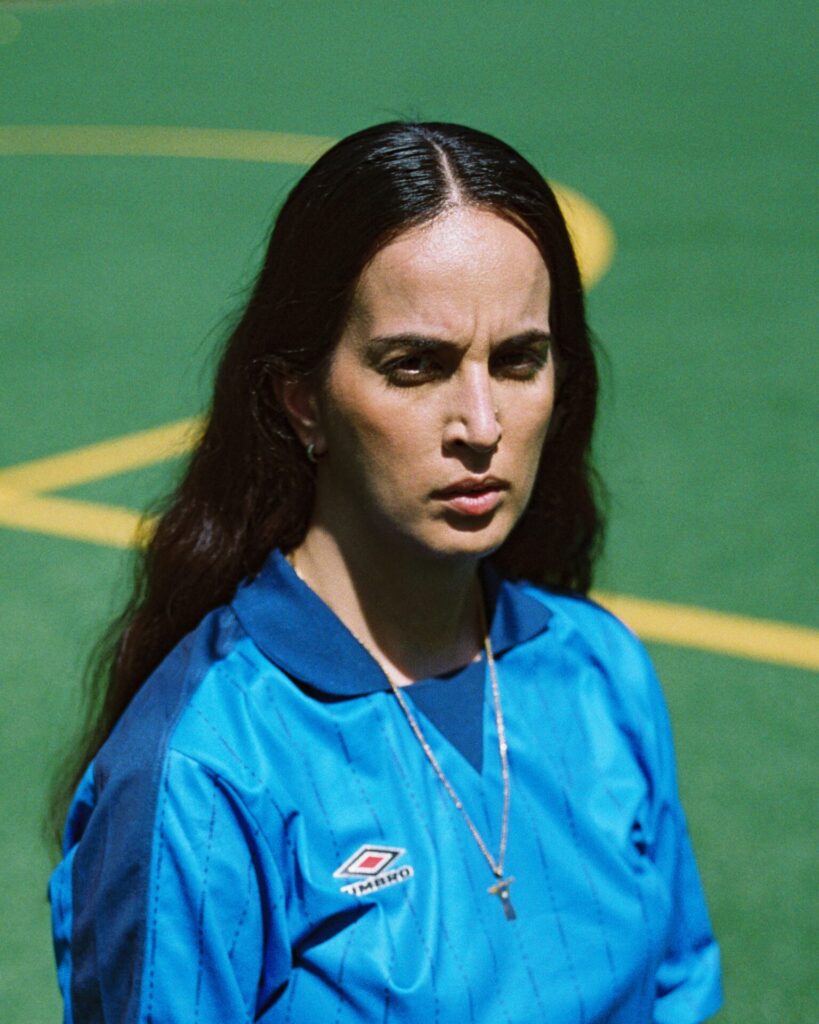 Frau mit langen dunklen Haaren trägt blaues Umbro-Sporttrikot und silberne Halskette mit Kreuzanhänger vor grünem Sportplatz mit gelben Linien.