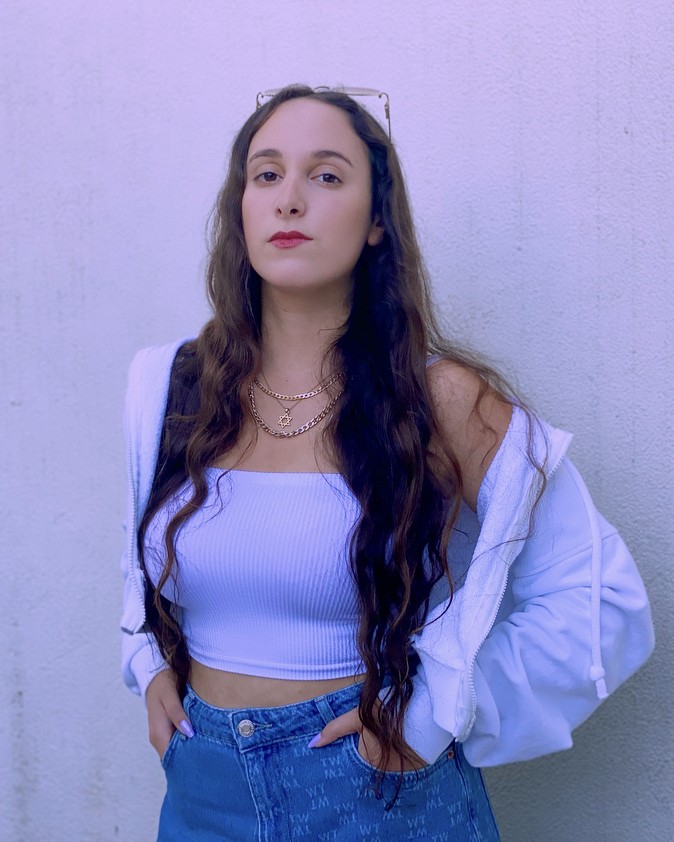 Frau mit langen, gewellten Haaren trägt weißes Crop-Top, blaue Jeans und weiße Jacke, steht vor hellvioletter Wand.