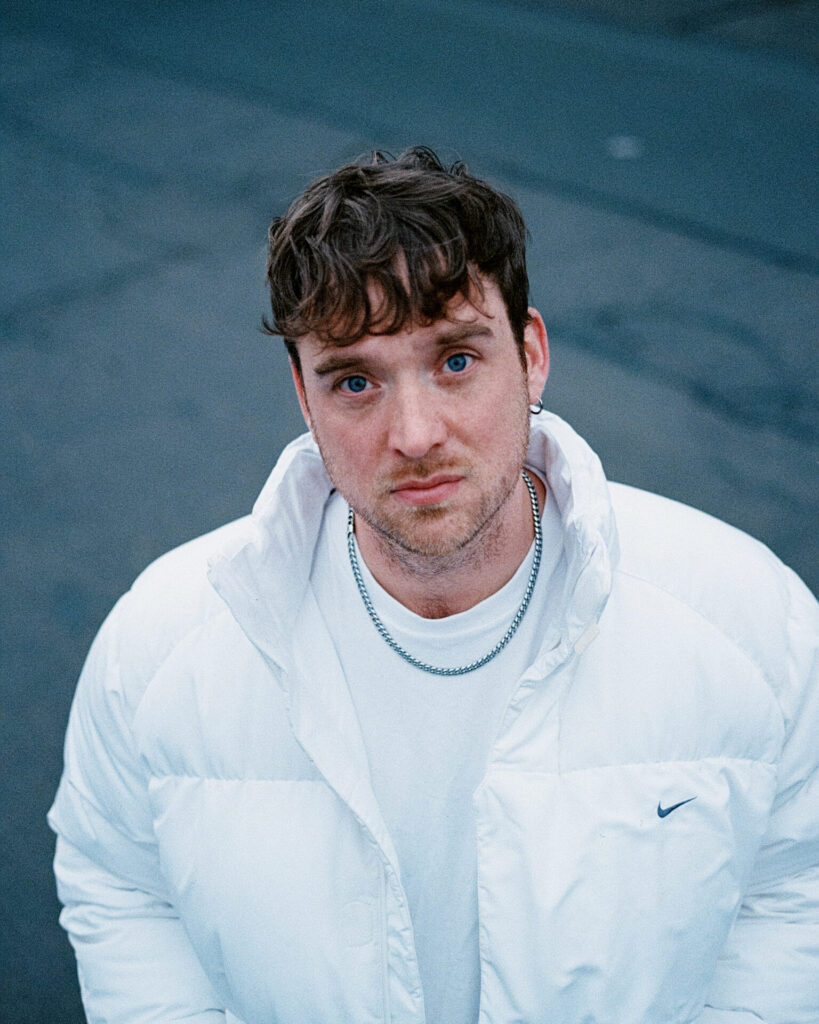 Person mit kurzem, gewelltem Haar trägt weiße Nike-Steppjacke, weißes T-Shirt und silberne Kette vor dunklem Hintergrund.
