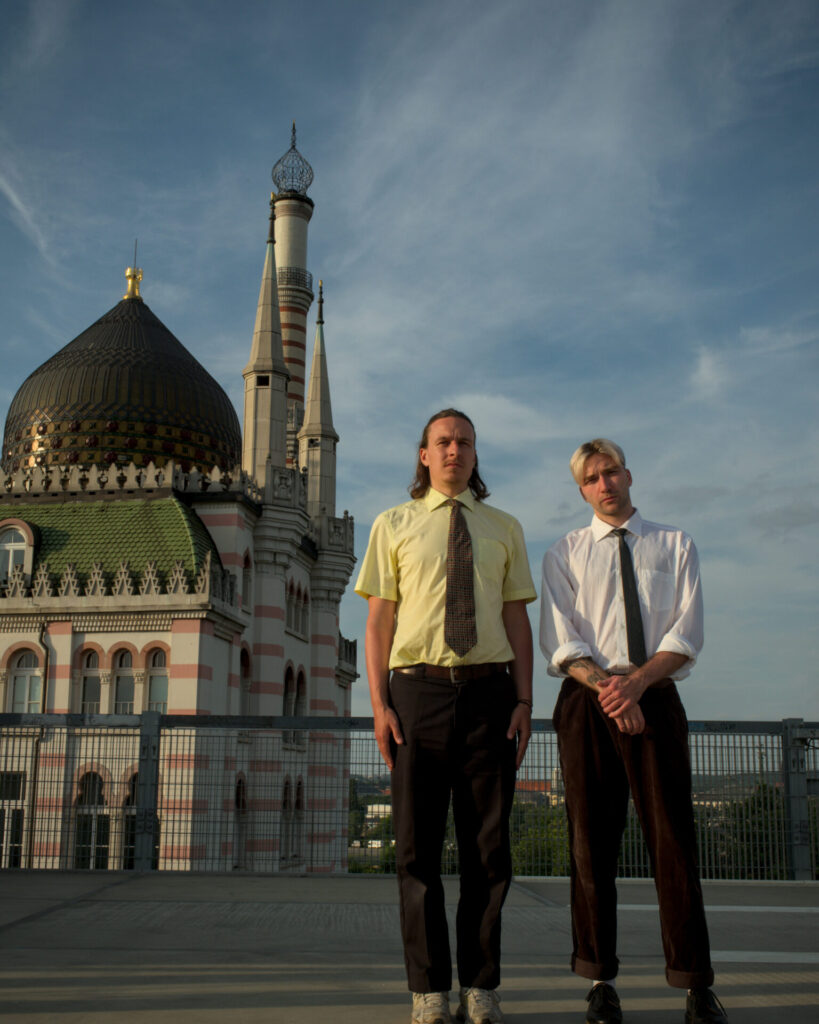 Zwei Männer in Hemd und Krawatte stehen vor einer historischen Moschee mit Kuppel und Minarett unter bewölktem Himmel.