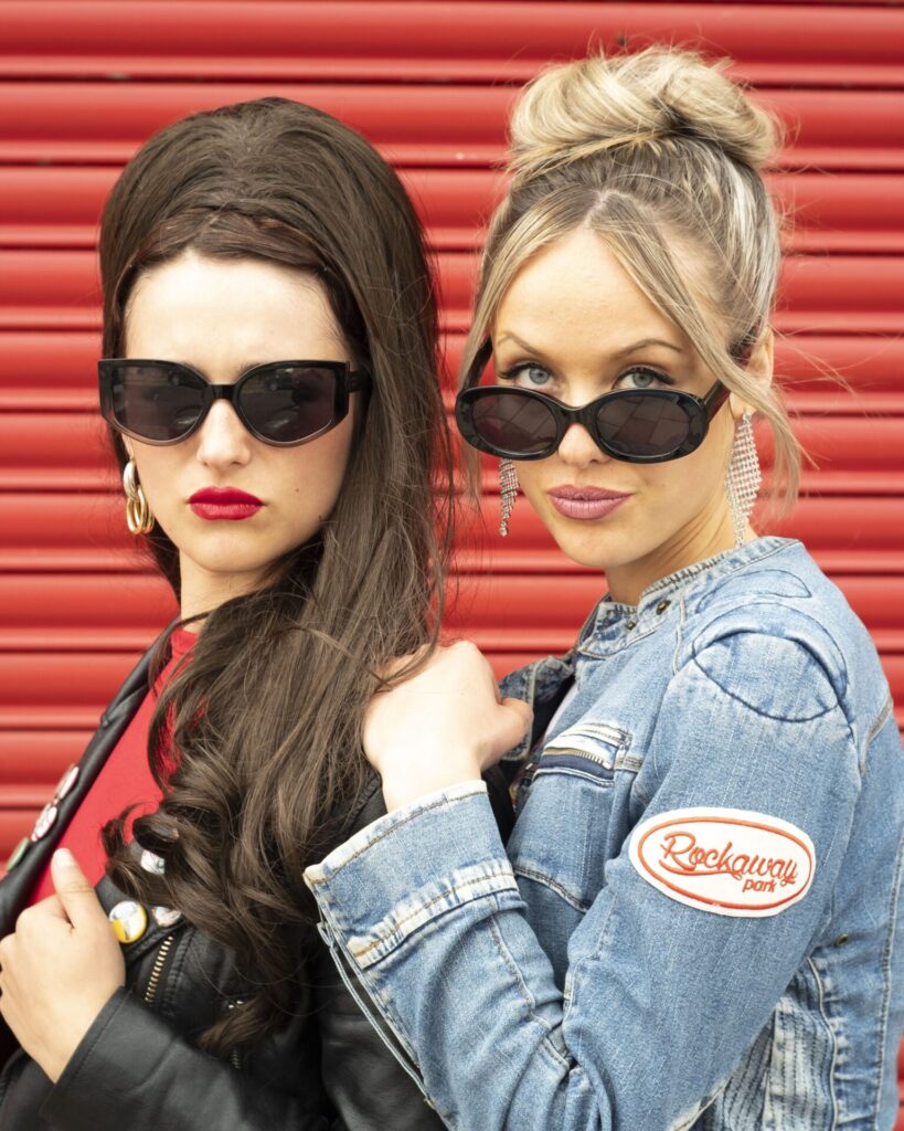 Zwei Frauen stehen vor einer roten Garagentür, eine trägt eine Jeansjacke mit einem "Rockaway park"-Aufnäher, die andere eine schwarze Lederjacke.
