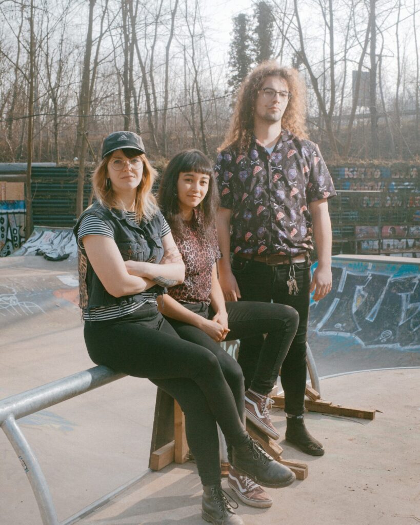 Drei Personen sitzen und stehen an einer Skateboardrampe im Freien mit Graffiti im Hintergrund