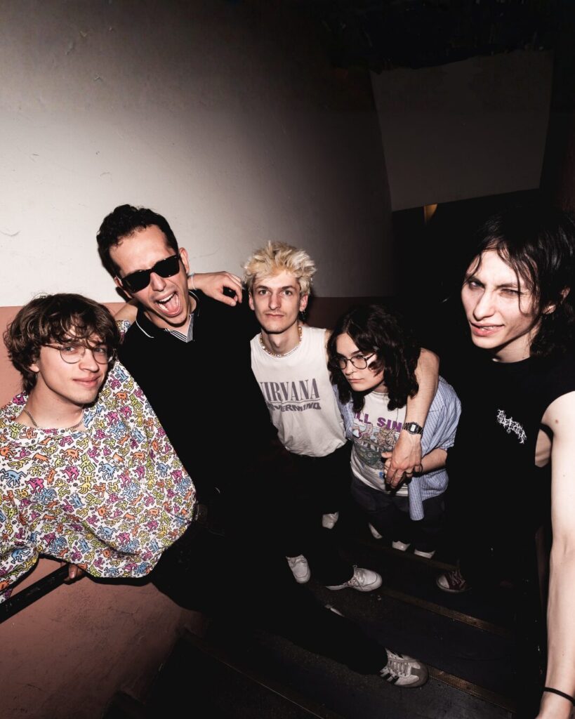 Gruppe von fünf Personen steht eng zusammen auf einer Treppe, eine Person trägt ein weißes Nirvana-T-Shirt, eine andere ein bunt gemustertes Oberteil.