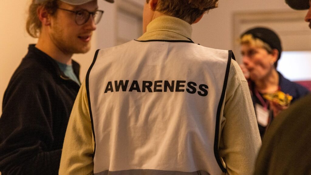 Person von hinten mit hellfarbener Weste und dem Schriftzug 'AWARENESS' auf dem Rücken in einer Gruppe von Menschen in Innenraum