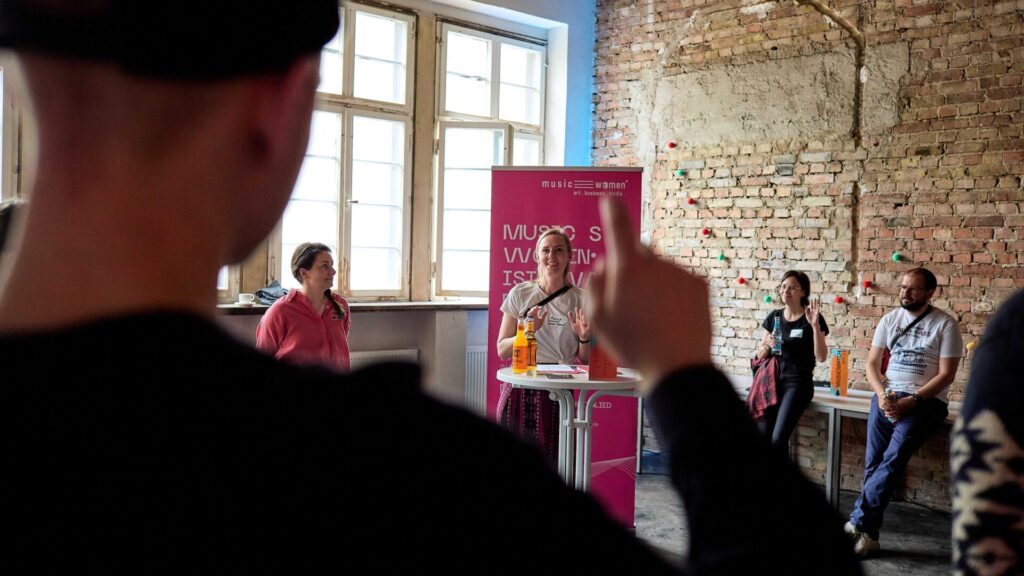 Person im Vordergrund mit erhobenem Zeigefinger vor einer Gruppe von vier Personen in einem Raum mit Backsteinwand und großen Fenstern