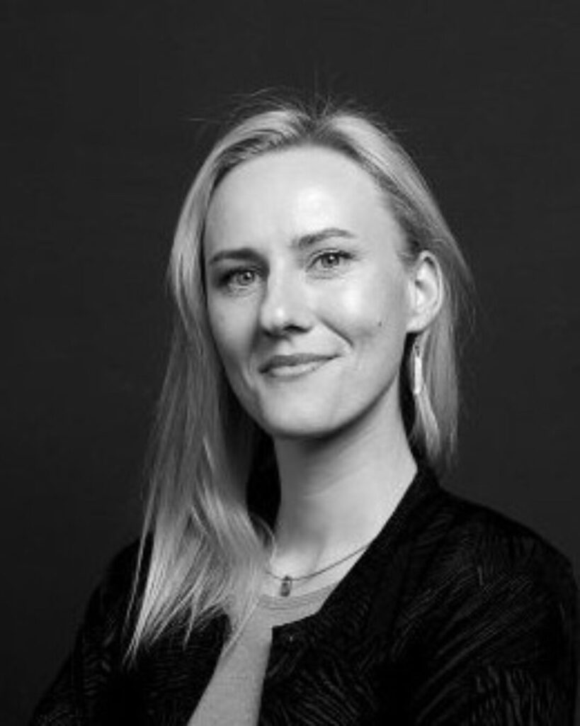 Eine Person mit langen Haaren, die eine Kette und Ohrringe trägt, vor dunklem Hintergrund