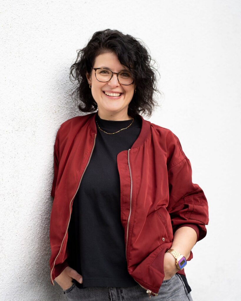 Person mit lockigem Haar lehnt an weißer Wand trägt rote Jacke, schwarzes Shirt, goldene Halskette und Armbanduhr mit blauem Zifferblatt