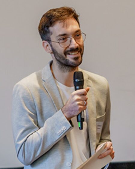 Mann mit Brille und grauem Sakko hält Mikrofon und Notizzettel in den Händen