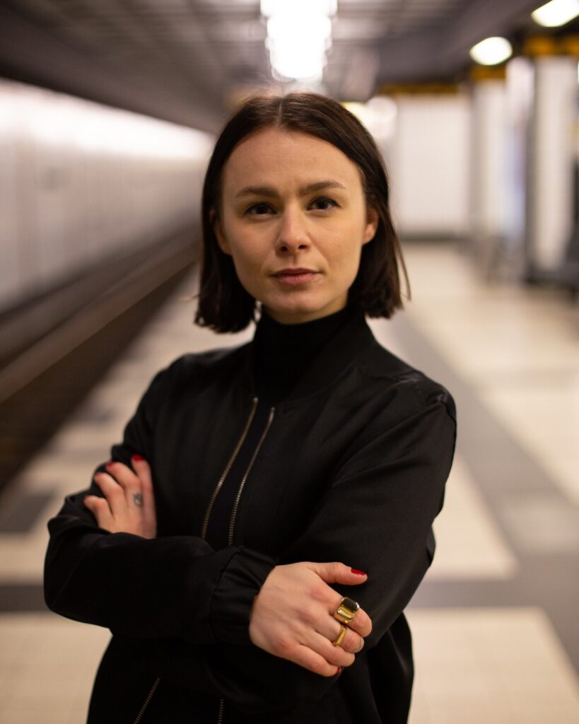 Person mit schulterlangem Haar und schwarzem Oberteil steht mit verschränkten Armen in einem U-Bahn Tunnel