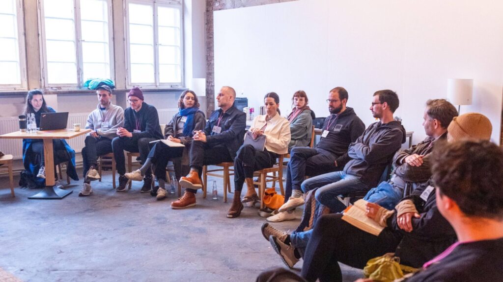 Gruppe von Menschen sitzt in einem Stuhlkreis in einem hellen Raum mit großen Fenstern und Backsteinwand