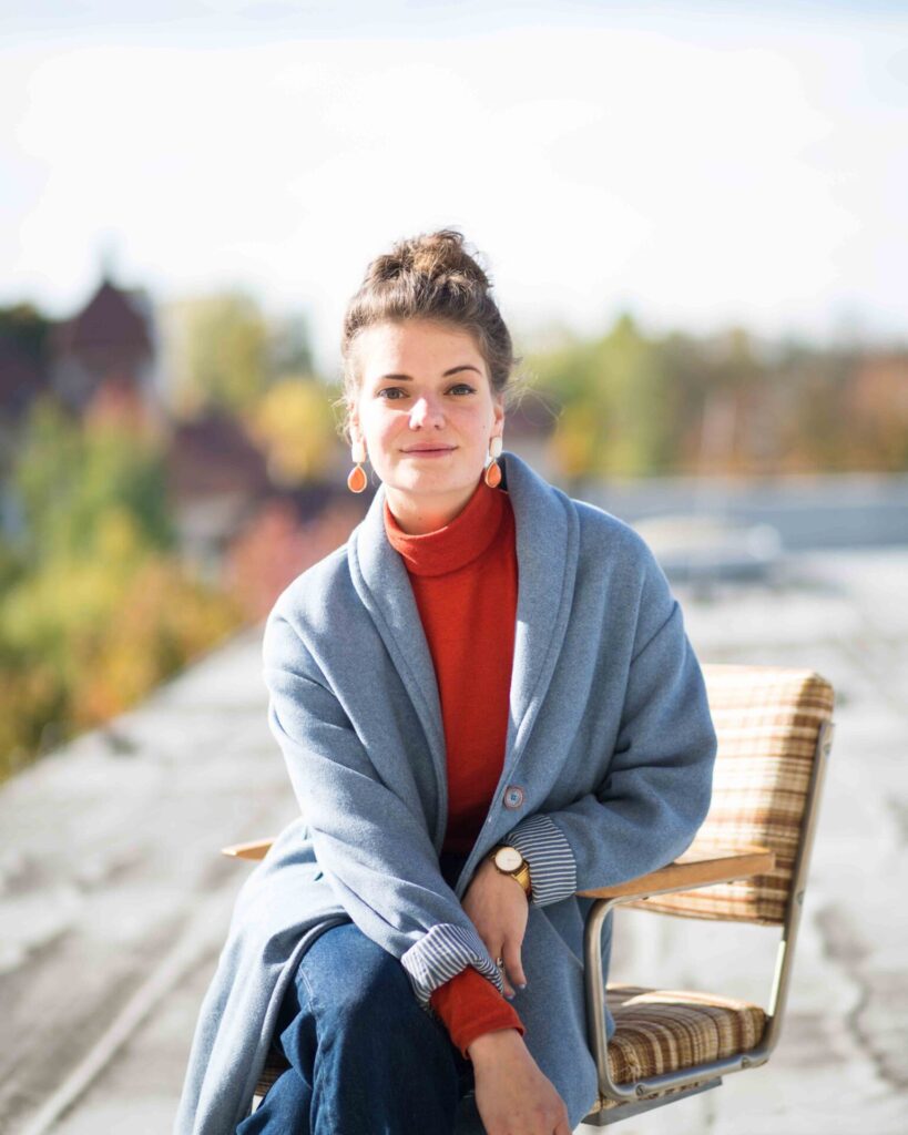 Person mit hochgestecktem Haar, rotem Rollkragenpullover und blauem Mantel sitzt auf einem Stuhl im Freien