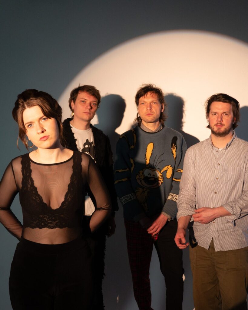 Vier Personen stehen vor einer hellen Wand mit rundem Lichtfleck, eine Frau trägt ein schwarzes, transparentes Oberteil mit Spitzenbesatz