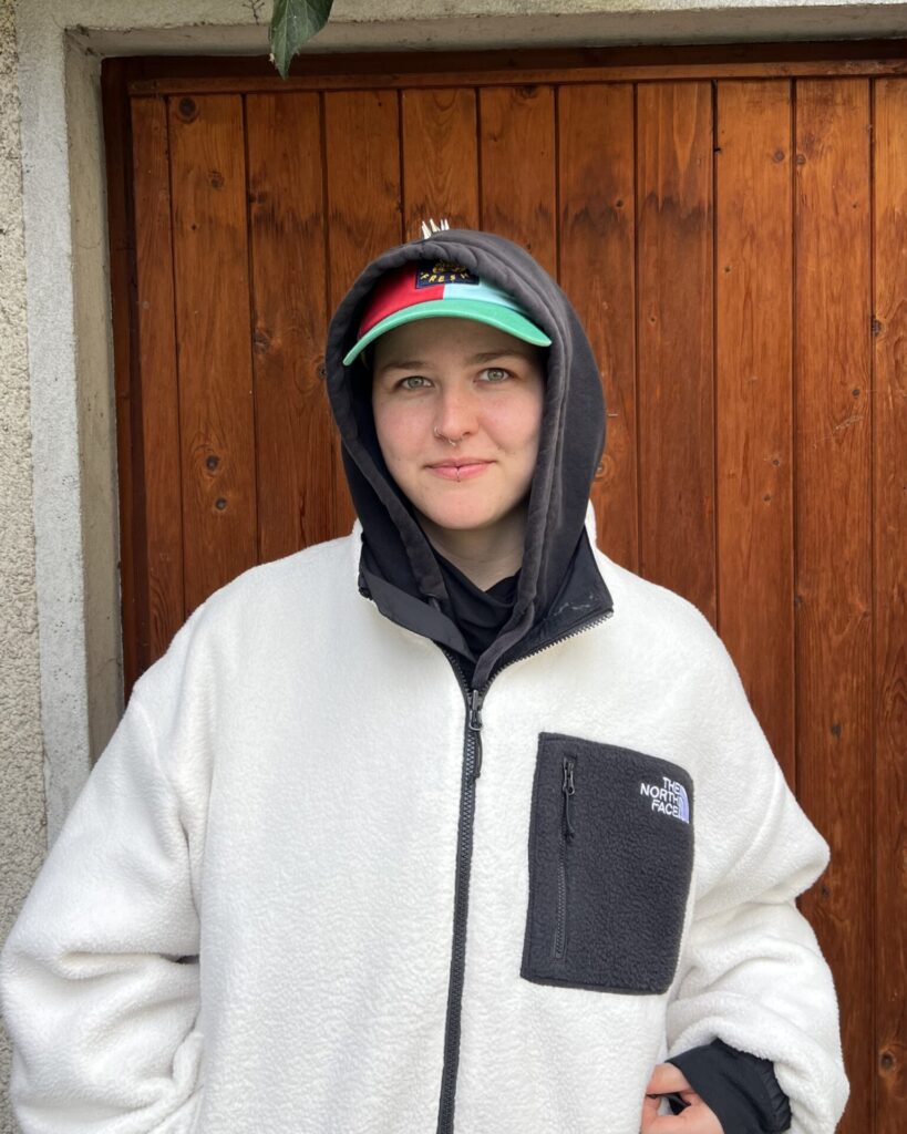 Person mit Kapuzenjacke und Baseballkappe steht vor einer Holztür
