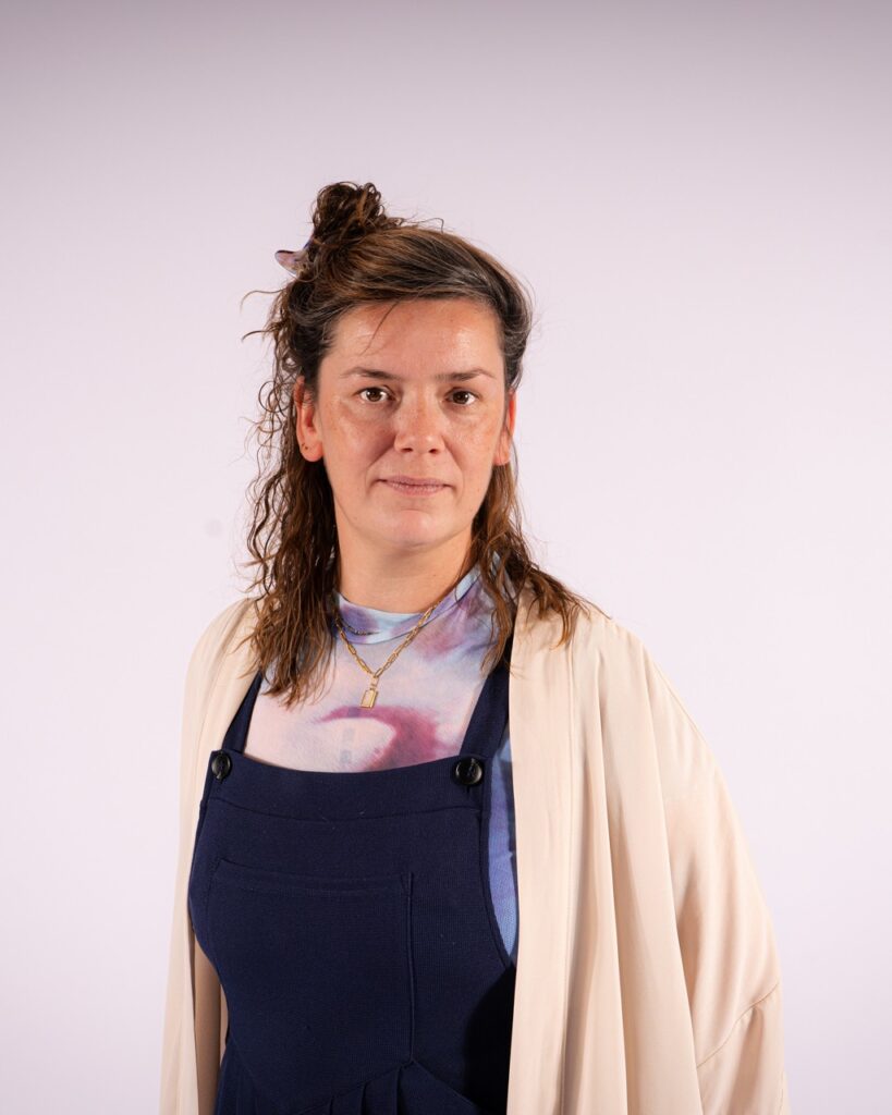 Person mit hochgebundenem lockigem Haar trägt ein helles, weites Oberteil über einer dunklen Latzhose und eine Kette mit Anhänger